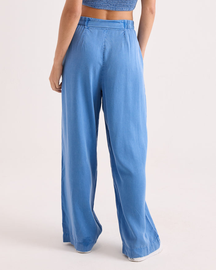 A person stands facing away, wearing Volcom Women’s Bow Chicka Pants—loose-fitting, cerulean wide-leg woven pants featuring an elastic waistband—paired with a blue cropped top. The background is plain and light-colored.