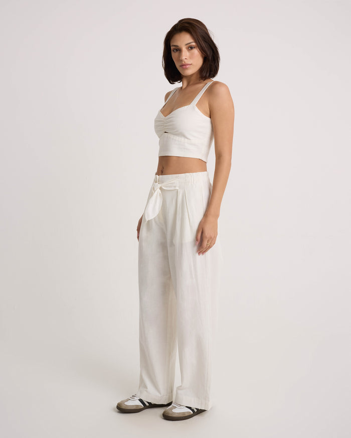 A woman with short dark hair poses against a plain light background, wearing Volcom's Women's Bow Chicka Pants—a high-waisted, wide-leg cotton-linen blend style with a tie-front waist—paired with a white crop top and white sneakers.