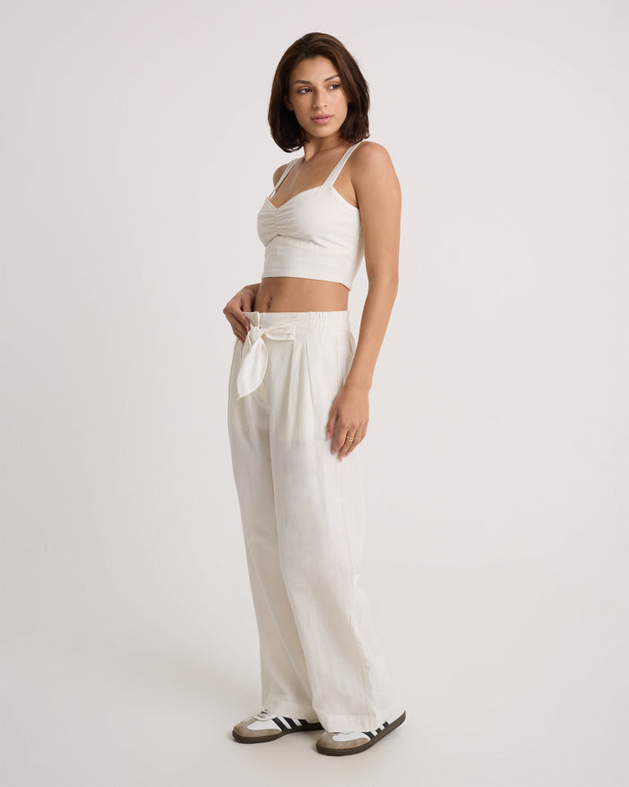 A woman models Volcom's Women's Bow Chicka Pants—loose, wide-leg, cotton-linen blend pants with a tie-front waist—paired with a white crop top and black and white sneakers against a plain background.