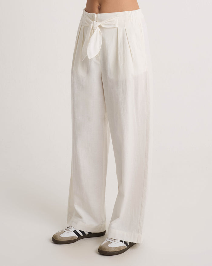 A person wears Volcom Women's Bow Chicka Pants, featuring a cotton-linen blend, wide legs, tie-front waist, and pleats, styled with black and white sneakers. The upper body is not visible against a light plain background.