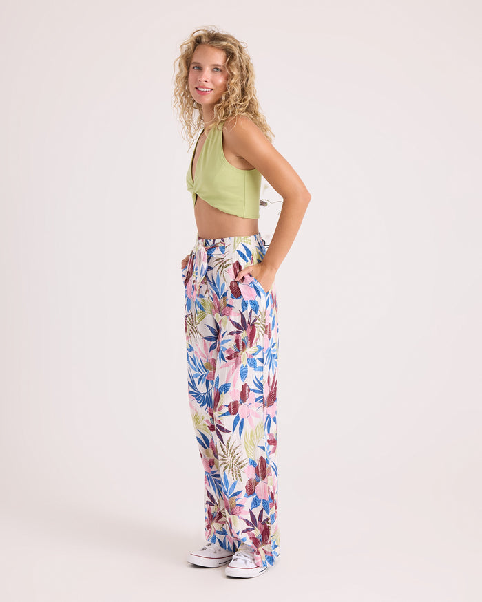 A woman with curly blonde hair wears a sleeveless green crop top, the Volcom Women's Bow Chicka Pants—colorful floral wide-leg pants featuring a relaxed fit and elastic waistband—and white sneakers. She stands sideways with her hands in her pockets against a plain light background.
