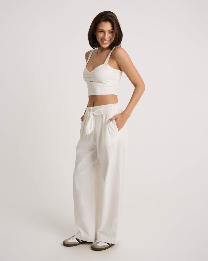A woman smiles in Volcom Women's Bow Chicka Pants, featuring a cotton-linen blend, wide legs, and a tie-front waist. She pairs them with a white crop top and sneakers, hands in pockets, posing against a plain white background.