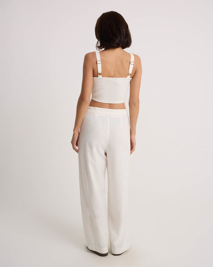 A person with shoulder-length dark hair faces away, wearing a white sleeveless crop top and Volcom Women's Bow Chicka Pants—loose, wide-leg cotton-linen blend trousers—against a plain light background.