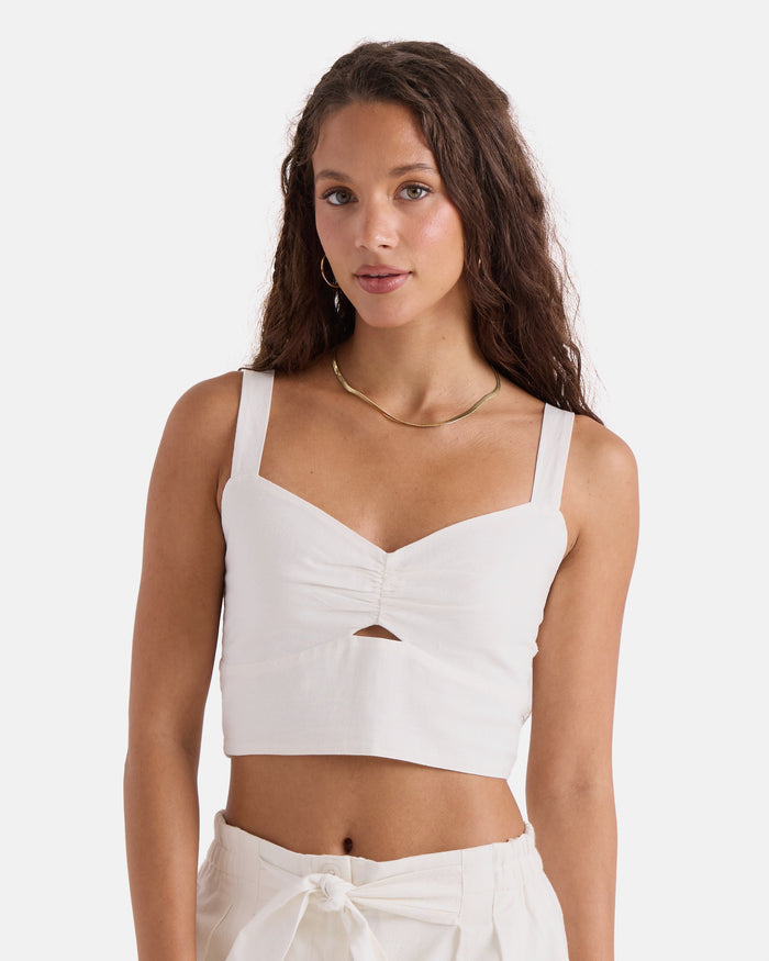 A woman with long, wavy brown hair models the Volcom Women's Bow Chicka Tank—a vintage white top with thick straps and a front cutout—paired with high-waisted white pants against a plain white background.