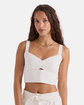 A woman with long, wavy brown hair models the Volcom Women's Bow Chicka Tank—a vintage white top with thick straps and a front cutout—paired with high-waisted white pants against a plain white background.