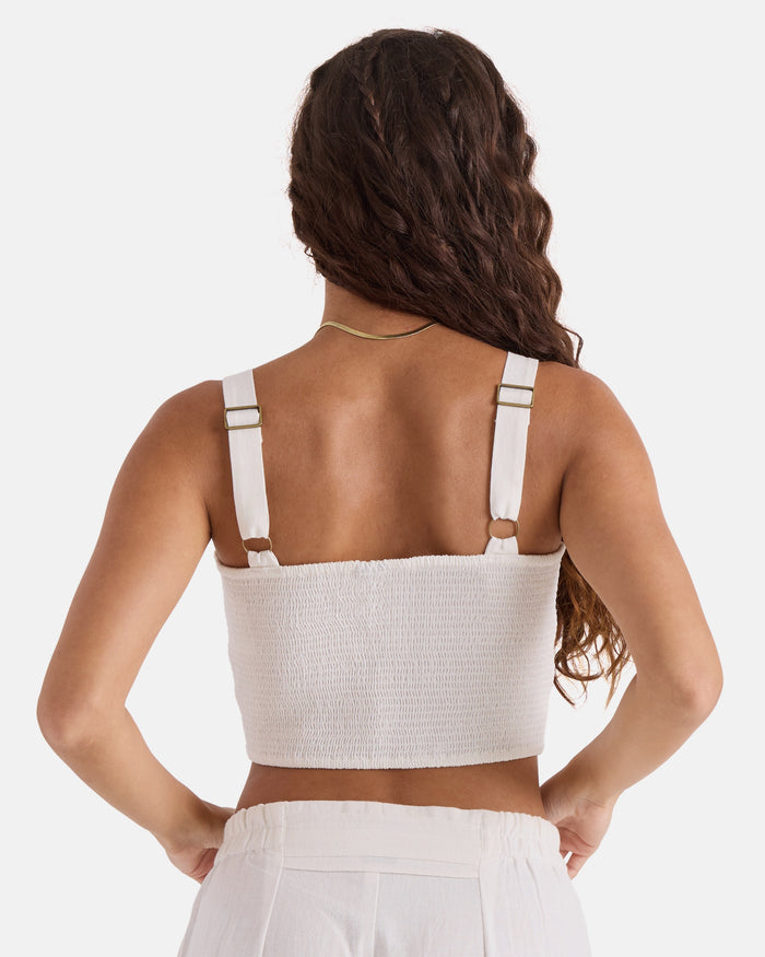 A woman with long wavy brown hair faces away, wearing the Volcom Women's Bow Chicka Tank— a vintage white crop top with adjustable straps—paired with high-waisted white cotton-linen blend pants against a plain light background.