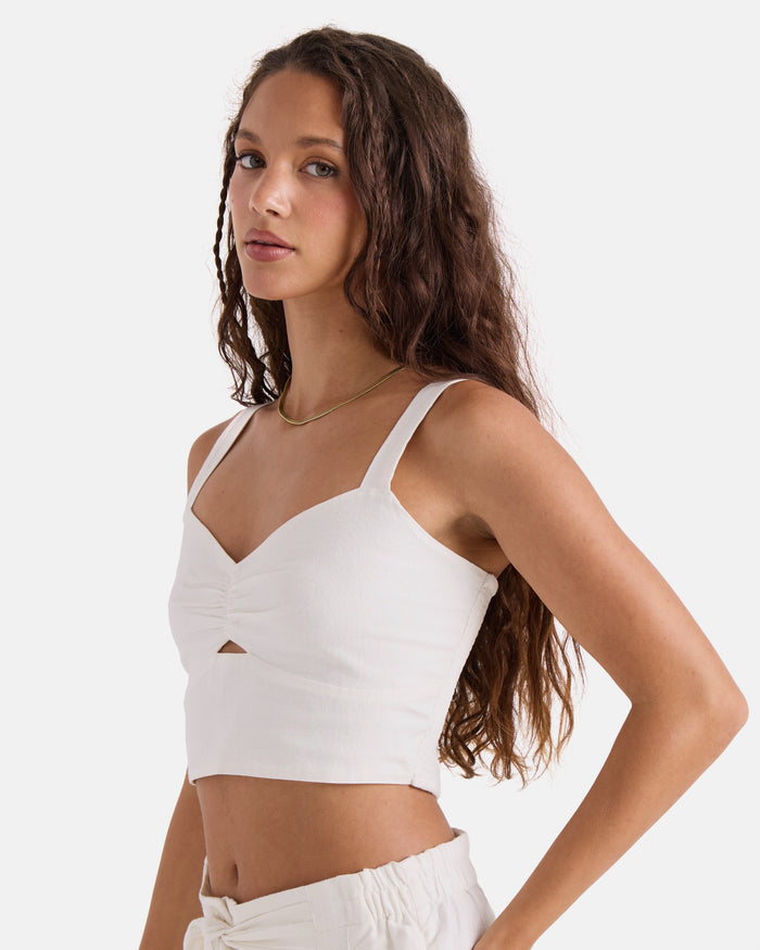 A woman with long wavy brown hair models the Volcom Women's Bow Chicka Tank in vintage white, paired with matching cotton-linen blend pants, standing sideways and looking at the camera against a plain white background.