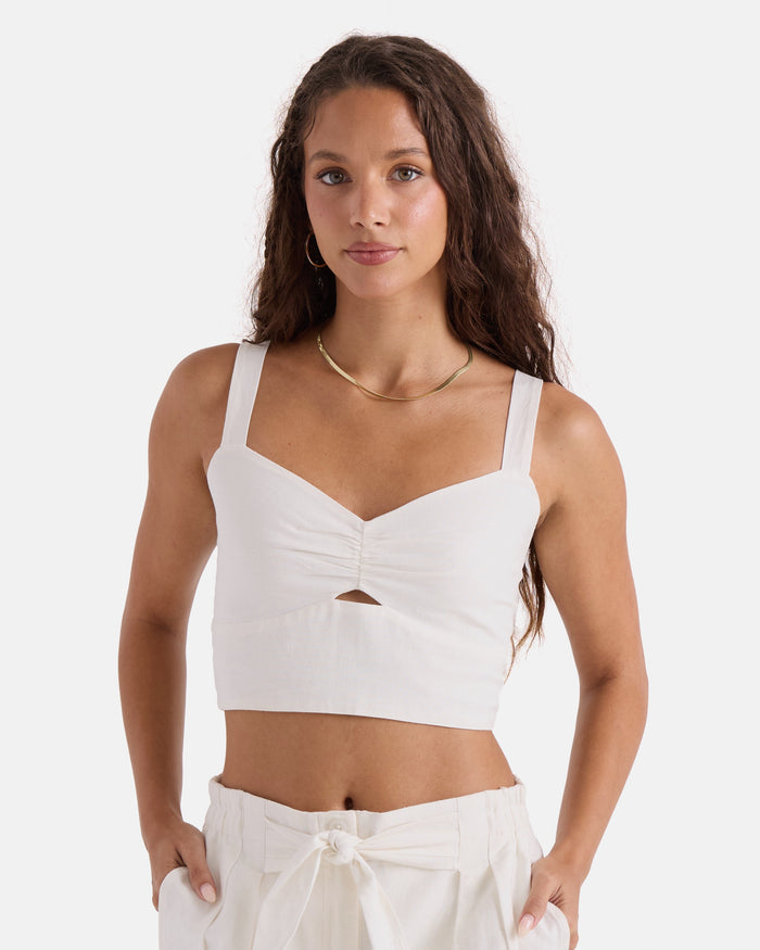 A woman with long wavy brown hair wears the Volcom Women's Bow Chicka Tank, a vintage white crop top featuring a front knot detail, paired with matching high-waisted pants. She stands with hands in pockets against a plain white background.