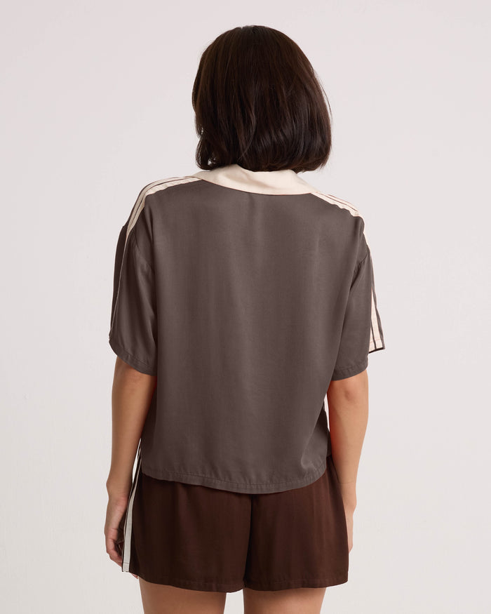 A woman with shoulder-length dark hair wears the Volcom Women's Boxing Shirt, a loose brown top with cream trim and a cropped silhouette, paired with matching brown shorts. She stands with her back to the camera against a plain, light background.