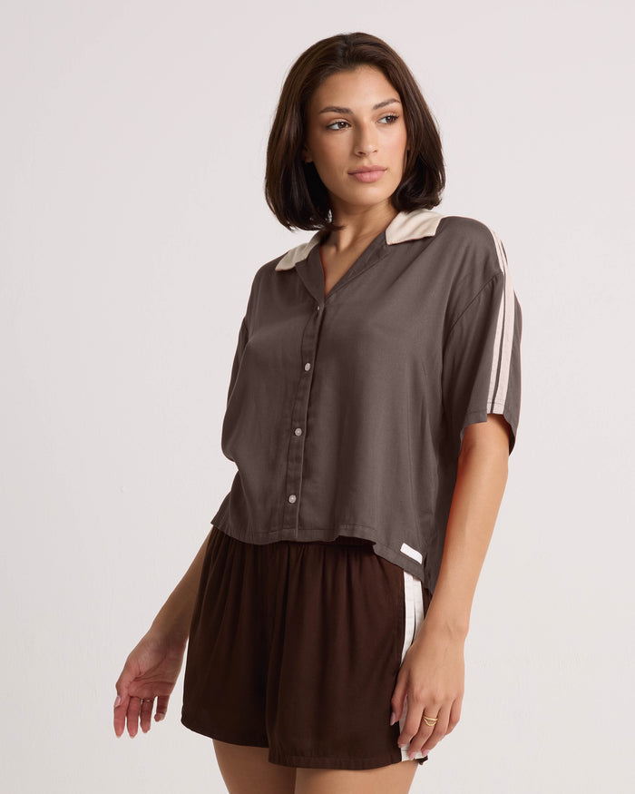 A woman wears the Volcom Women's Boxing Shirt, a loose brown top with short sleeves, cream details, and a cropped silhouette, paired with matching brown shorts featuring a cream stripe, standing against a plain background.