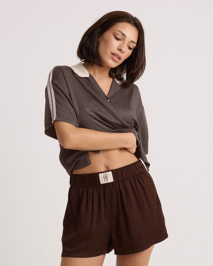 A woman with shoulder-length brown hair wears the Volcom Women's Boxing Shirt, featuring a loose, cropped silhouette, paired with matching brown shorts. She stands against a plain white background, lifting her shirt slightly with one hand.