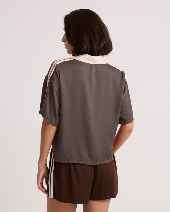 A woman with short, dark hair faces away from the camera, modeling a loose-fitting cropped Volcom Women's Boxing Shirt in brown with light shoulder stripes, paired with matching brown shorts featuring white side stripes.