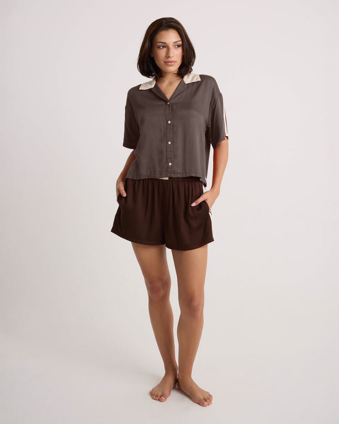 A woman with short dark hair is barefoot, standing against a plain white background with her hands in her pockets, wearing the Volcom Women's Boxing Shirt—featuring a cropped brown button-up design with a light collar—and matching brown shorts, looking slightly to the side.