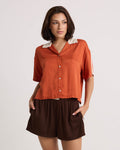 A woman with short brown hair stands against a plain background, looking to the side with one hand in her pocket. She wears a lightweight orange collared short-sleeve shirt paired with Volcom Women's Boxing Shorts—brown shorts featuring an elastic waistband.