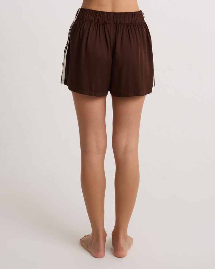A person stands barefoot on a white surface, shown from the back, wearing Volcom Women's Boxing Shorts—lightweight brown shorts with an elastic waistband and white side stripes.