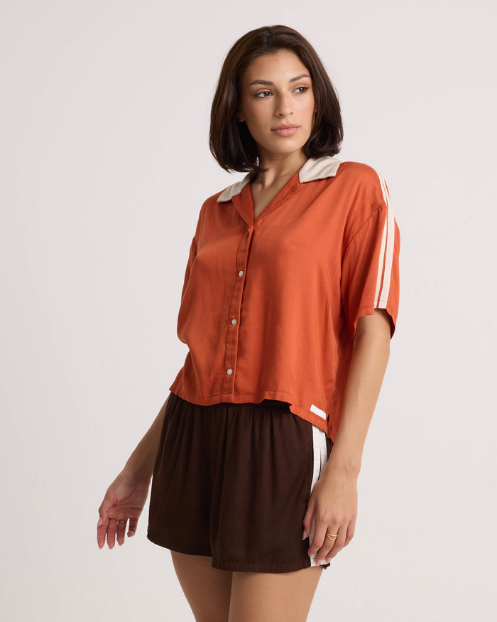 A woman stands against a plain light background wearing Volcom Women's Boxing Shorts—lightweight brown shorts with an elastic waistband—and an orange button-up short-sleeve shirt featuring white stripes on the sleeves.