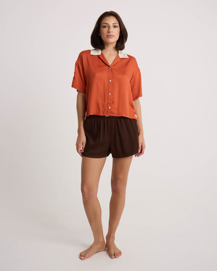 A woman stands barefoot against a plain background, wearing a lightweight, short-sleeved orange button-up shirt with a white collar and loose brown Volcom Women's Boxing Shorts featuring an elastic waistband.