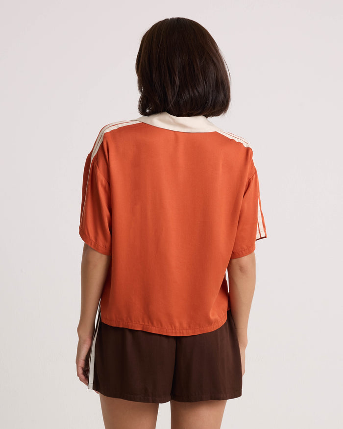 A person with shoulder-length dark hair faces away, wearing an orange short-sleeve top with white stripes on the sleeves and lightweight brown Volcom Women's Boxing Shorts featuring an elastic waistband. The background is plain and light-colored.