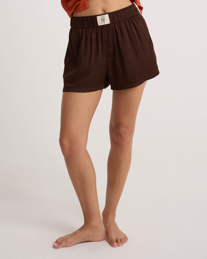 A person is standing barefoot against a plain light background, wearing Volcom Women’s Boxing Shorts in lightweight dark brown fabric, featuring an elastic waistband and a small white patch on the front.