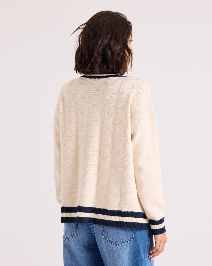 A woman with short brown hair, dressed in the Volcom Women's Boyfriend Cardigan in cream with black striped cuffs and collar, dropped shoulders, and blue jeans, stands facing away from the camera against a light background.