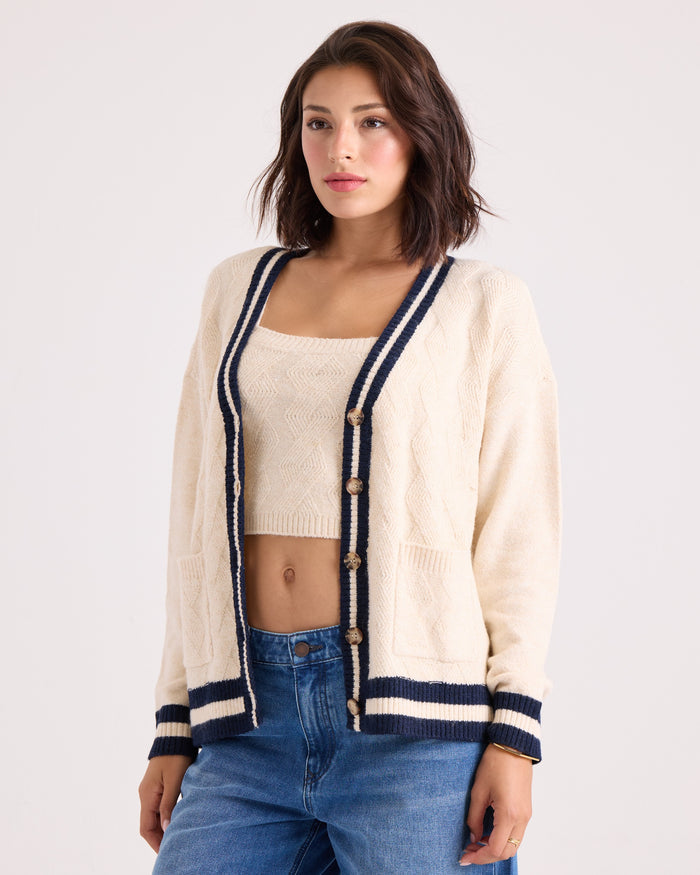 A woman stands against a plain light background, looking slightly to the side, dressed in the Volcom Women's Boyfriend Cardigan—a cream knit design with navy trim, dropped shoulders, and patch pockets—layered over a matching crop top and paired with blue jeans.