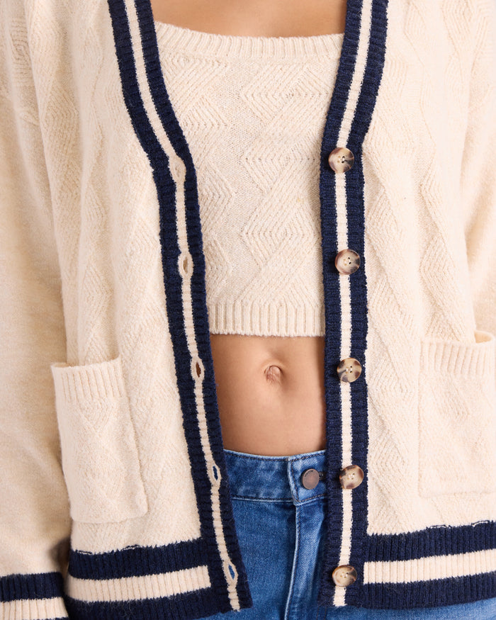 A person wearing the Volcom Women's Boyfriend Cardigan in cream with navy trim, buttons, and patch pockets over a matching cream crop top and blue jeans. The cardigan has dropped shoulders for a relaxed fit. Only the torso is visible.