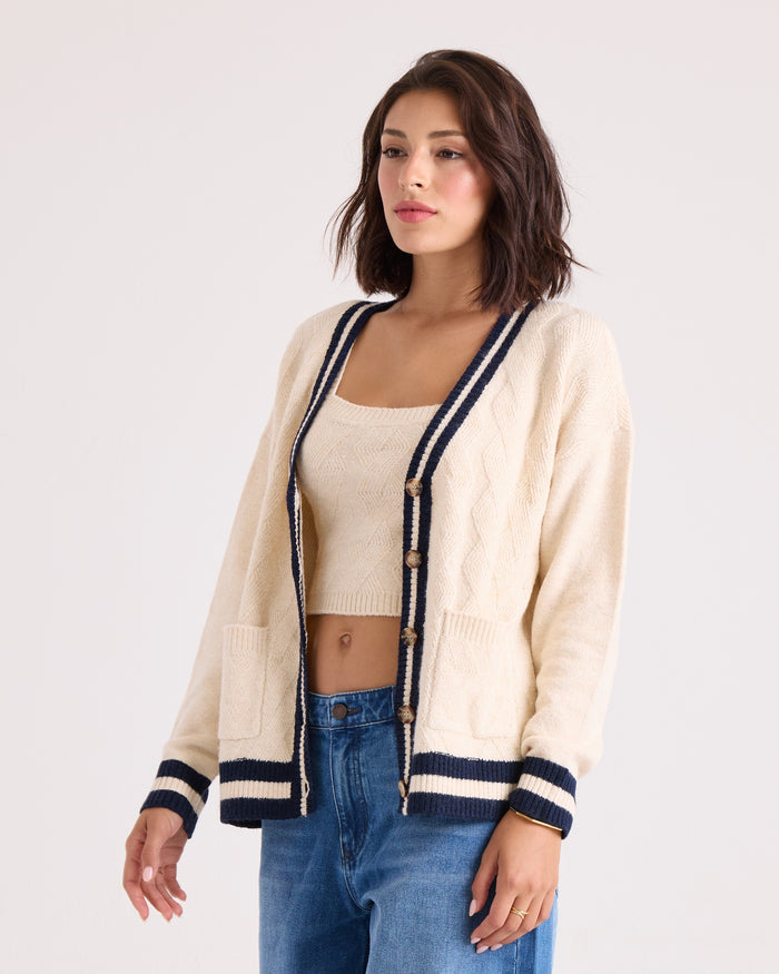 A woman stands against a plain light background wearing the Volcom Women's Boyfriend Cardigan in cream with navy trim, detailed with patch pockets and dropped shoulders, layered over a matching crop top and paired with blue jeans.