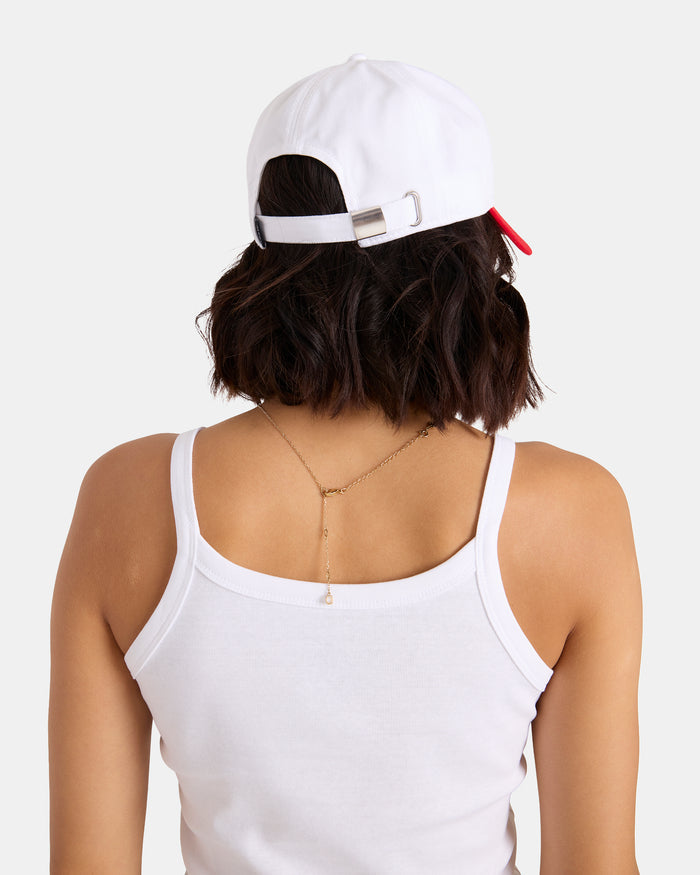 A person with short brown hair wears a white tank top and the Volcom Women's Breakin Hearts Hat, a white structured twill cap with an adjustable back strap, shown from behind against a plain background. A delicate chain necklace drapes down their back.