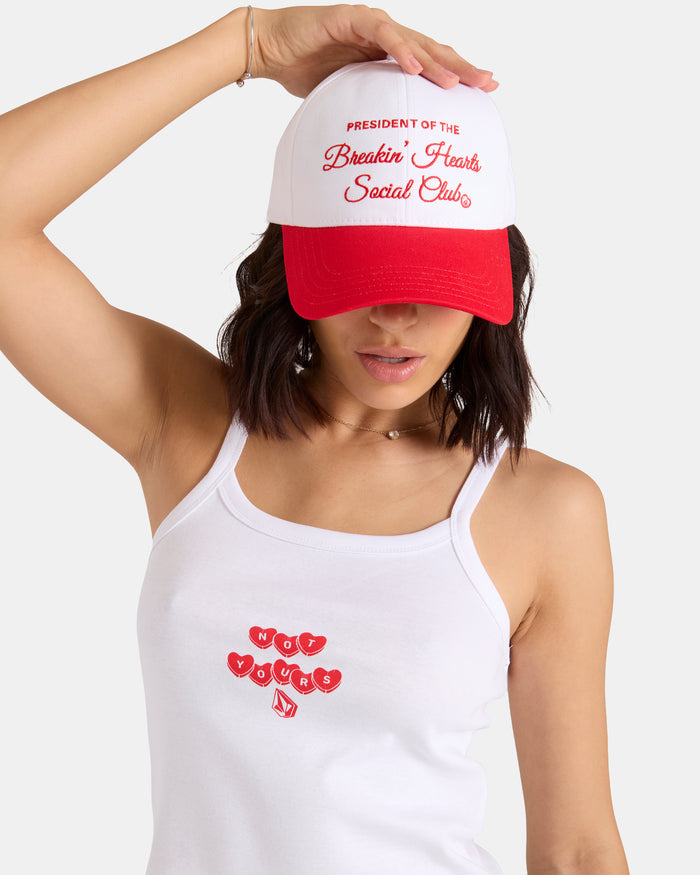 A woman tilts her Women's Breakin Hearts Hat by Volcom, a white structured twill cap with "President of the Breakin’ Hearts Social Club" embroidered on the front. She pairs it with a white tank top reading "Not Yours" in red text and hearts.