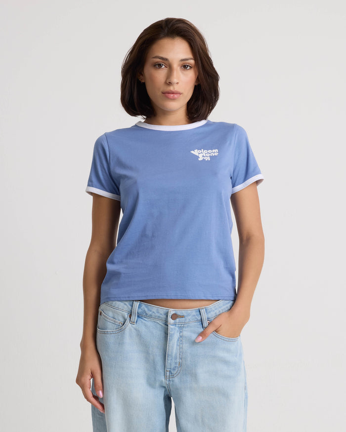A woman with short brown hair wears the Volcom Women's Brk Up Ringer Tee in Horizon Blue with white trim and a classic crew neck, paired with faded blue jeans, standing against a plain white background with one hand in her pocket.