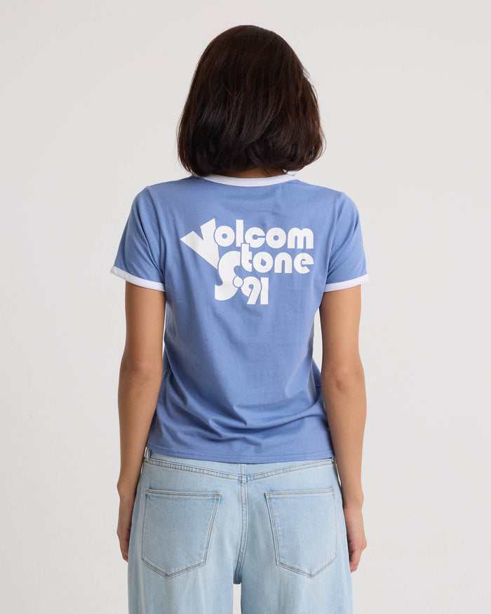 A person with shoulder-length dark hair, seen from behind, wears the Volcom Women's Brk Up Ringer Tee in Horizon Blue with a crew neck and "Volcom Stone S91" in white on the back, paired with light blue jeans.