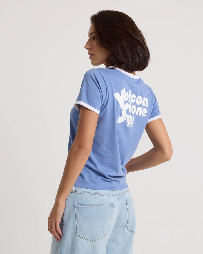 A woman stands sideways, looking left, wearing the Volcom Women's Brk Up Ringer Tee in Horizon Blue with a crew neck and "Volcom Stone 91" on the back, paired with light blue jeans against a plain light background.