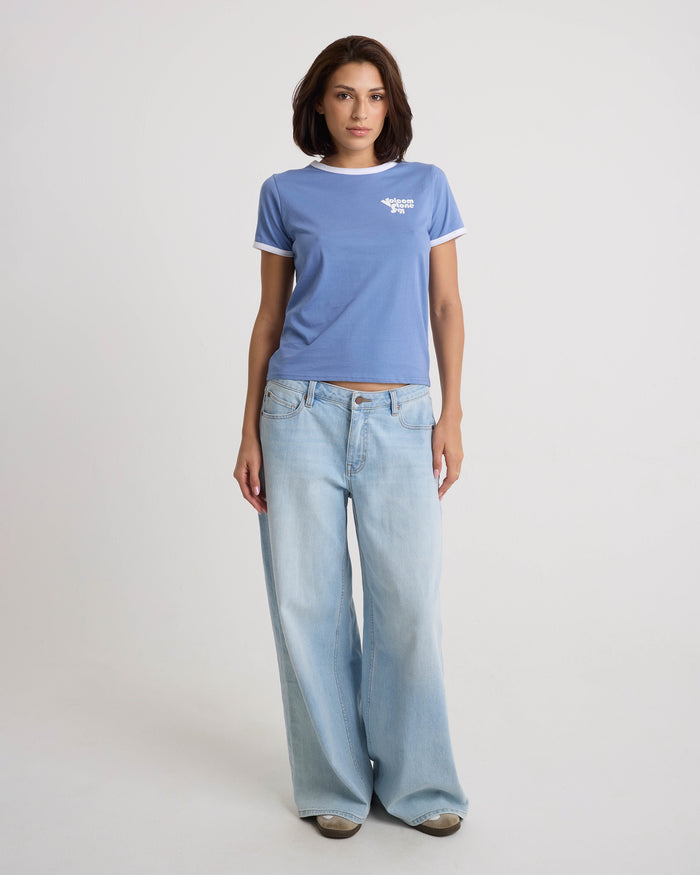 A woman faces forward with a neutral expression, wearing the Volcom Women's Brk Up Ringer Tee in Horizon Blue, paired with loose light blue jeans and sneakers against a plain white background.