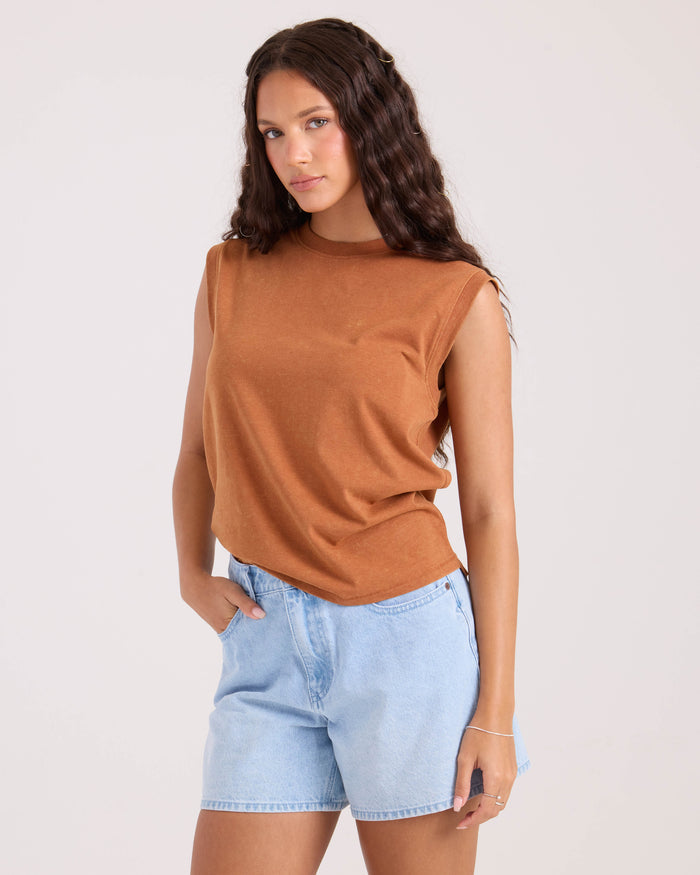 A woman with long wavy brown hair wears the Volcom Women's Burly sleeveless brown top and light blue denim shorts, standing against a plain light background with one hand in her pocket.