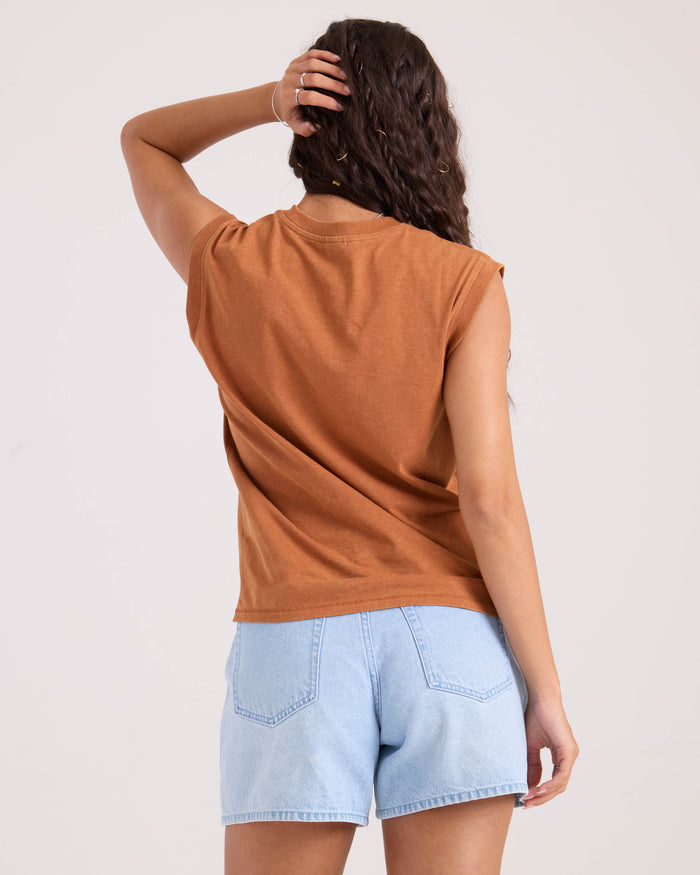 A woman with long wavy hair stands facing away, wearing the Volcom Women's Burly brown sleeveless top and light blue denim shorts. One hand is raised to the back of her head, set against a plain white background.
