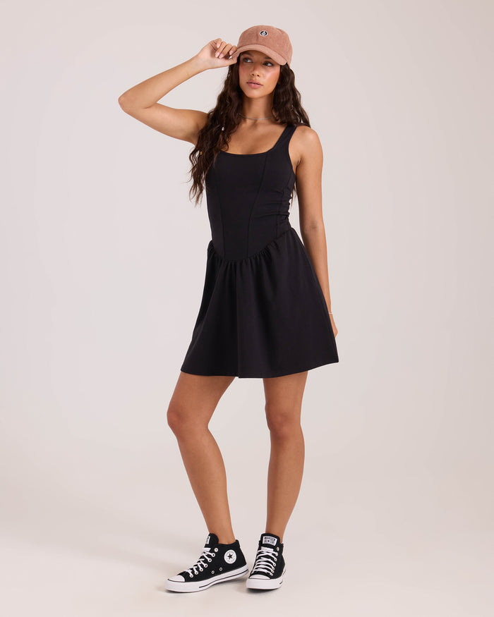A woman poses against a plain background, wearing the Volcom Women's Carve dress, black high-top Converse sneakers, and a light brown baseball cap. She touches the brim of her cap with one hand.