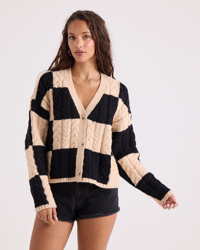 A woman wears the Volcom Women's Check You Sweater, a beige and black checkered button-front knit cardigan, paired with black shorts. She stands against a plain white background, gazing slightly to the side in a relaxed pose that highlights the comfy and effortless style of this piece.