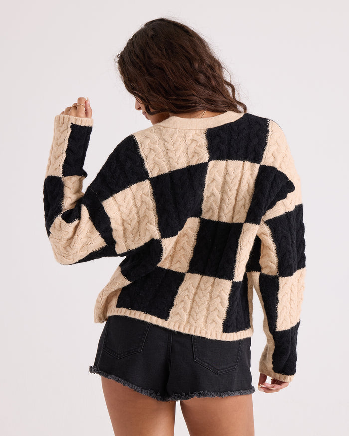 A person with long wavy hair, seen from behind, wears the Volcom Women's Check You Sweater—a comfy knit black and beige checkered cable-knit V-neck—paired with black denim shorts against a plain light background.
