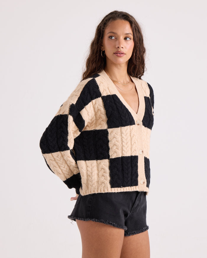 A woman is wearing the Volcom Women's Check You Sweater, a beige and black checkered knit button-front cardigan, paired with black shorts. She stands against a plain white background and looks to the side.