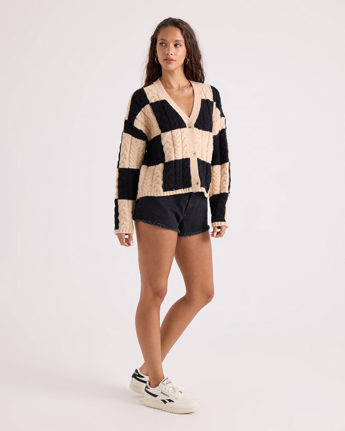 A woman with long wavy hair models the Volcom Women's Check You Sweater—a beige and black checkerboard patterned V-neck cardigan with a comfy knit and button front—paired with black denim shorts and white sneakers, standing against a plain white background.