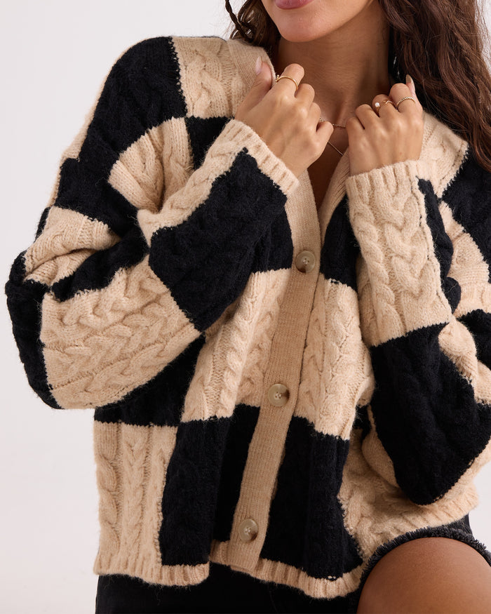A woman wearing the Volcom Women's Check You Sweater—a comfy, chunky knit cardigan featuring a large black and beige checkered pattern, cable knit texture, and a pearl button front—holds the collar with both hands.