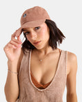 A woman with short dark hair wears the Volcom Women's Circle Stone Cord Hat, a sleeveless beige top, a simple necklace, and a bracelet. She looks at the camera while holding the brim of her hat.
