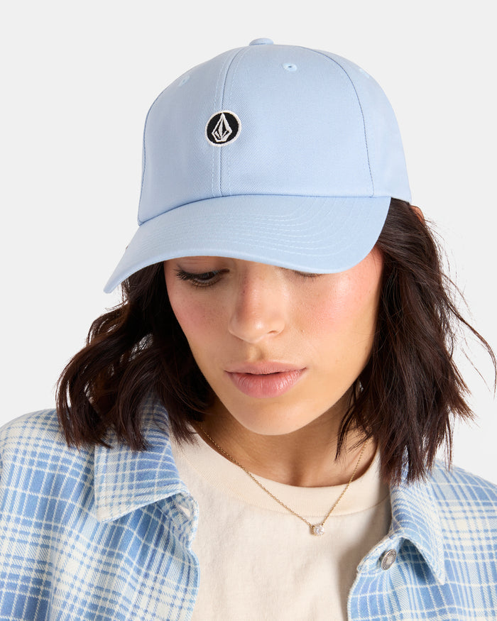 A woman wearing the Volcom Women's Circle Stone Dad Hat in light blue, paired with a plaid blue and white shirt, cream top, and delicate necklace, looks downward against a plain background.
