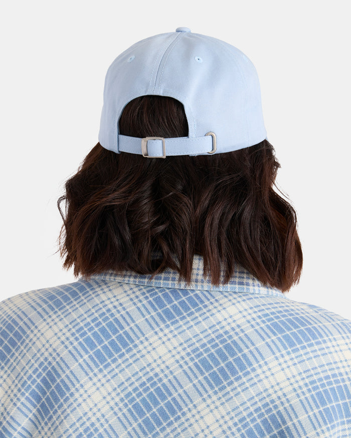 A woman with shoulder-length dark hair is seen from behind against a plain background, wearing Volcom’s Women’s Circle Stone Dad Hat in light blue and a blue-and-white plaid shirt.