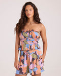 A woman with long wavy hair models the Volcom Women's Coastal Girly Cami with adjustable straps and matching shorts, both in a vibrant pink, purple, blue, and tan abstract pattern. She stands against a plain light background.