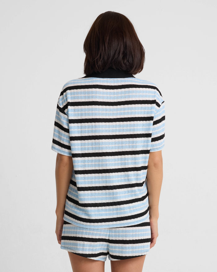 A person with shoulder-length dark hair stands with their back to the camera, wearing Volcom's Women's Coco Ho Crochet Shorts in light blue, white, and black stripes. The set has relaxed fit short sleeves. The background is plain white.