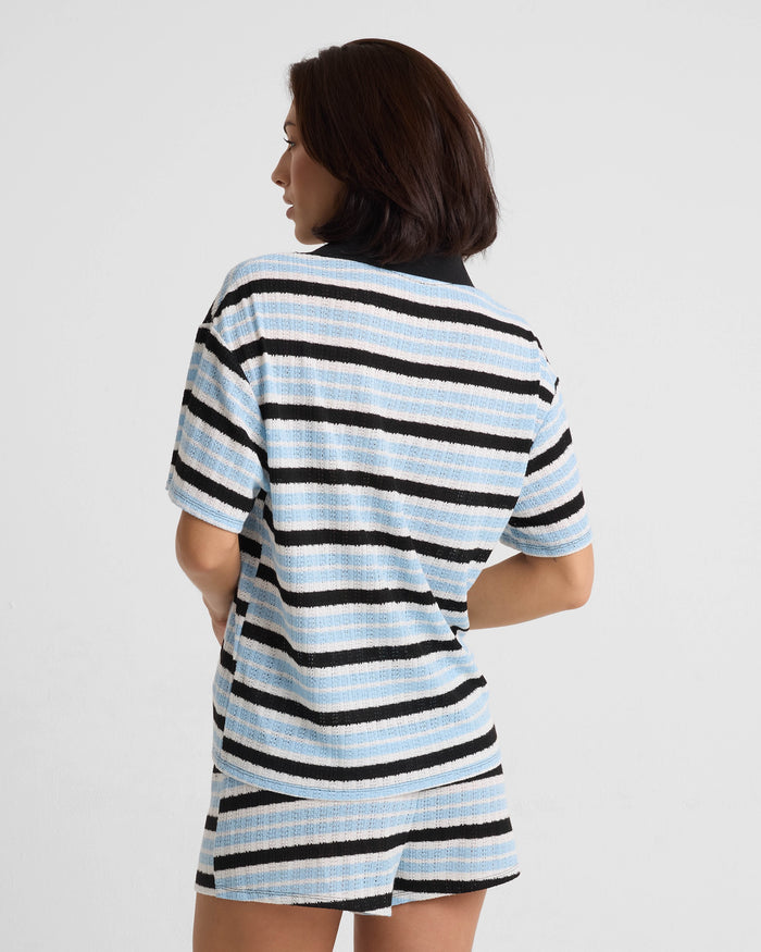 A woman with short dark hair stands facing away, wearing Volcom's Women's Coco Ho Crochet Shorts and a matching relaxed-fit short-sleeve shirt with horizontal blue, black, and white stripes against a plain light background.