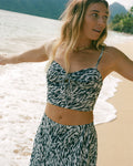 A woman with long, wavy hair stands on a sandy beach near the shoreline, arms outstretched, wearing the Volcom Women's Coco Ho Money Mama Cami. The ocean and lush green mountains are visible in the background.