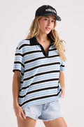 A person wears a relaxed fit Volcom Women's Coco Ho Rugby Top in blue, black, and white stripes, paired with a black baseball cap and light denim shorts against a plain background.