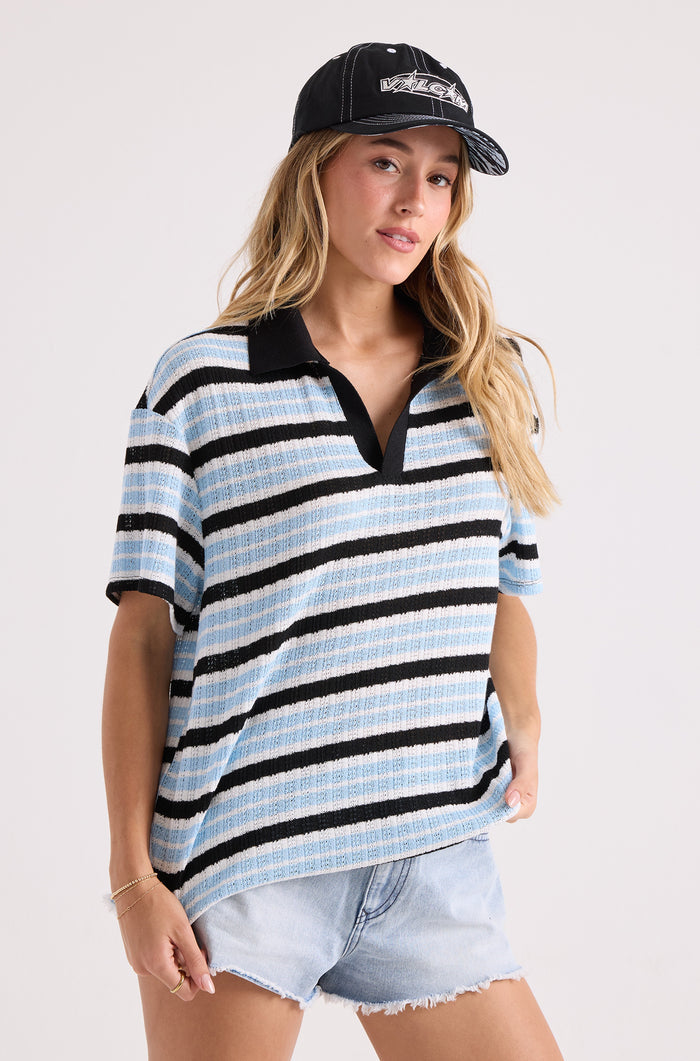 A person wears a relaxed fit Volcom Women's Coco Ho Rugby Top in blue, black, and white stripes, paired with a black baseball cap and light denim shorts against a plain background.